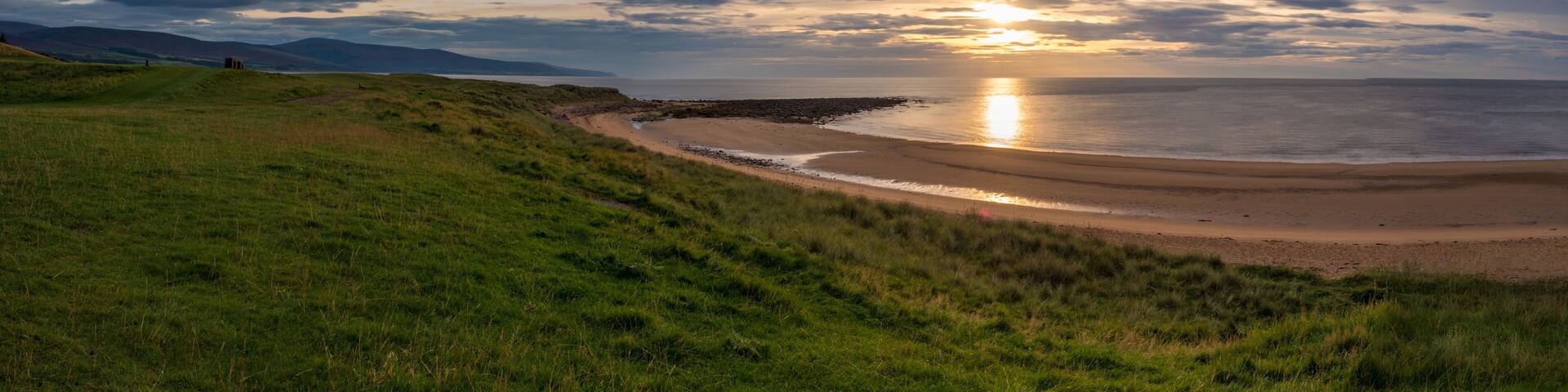 A coastal landscape with a grassy hill, a rocky shoreline, and a sunset over the ocean.