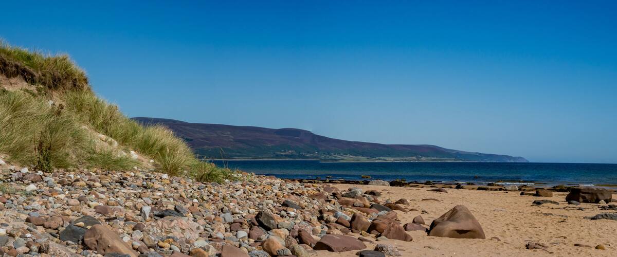 A rocky beach with a grassy slope and a view of the sea and distant hills.