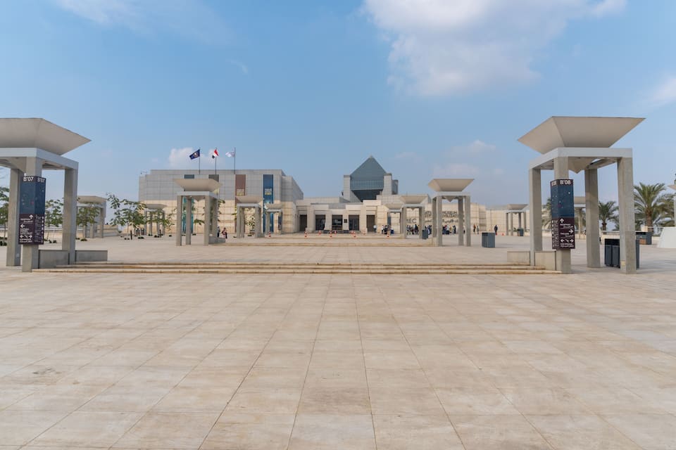 A large open plaza with a modern building in the background, featuring a prominent glass pyramid structure.
