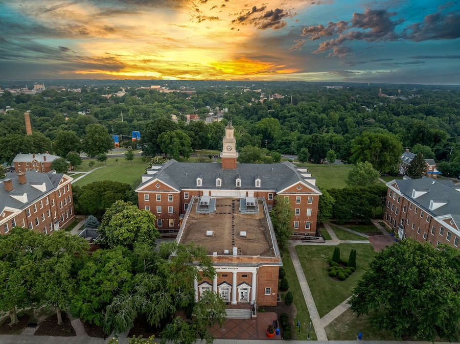 A large brick building with a clock tower, surrounded by green trees and a sunset sky.