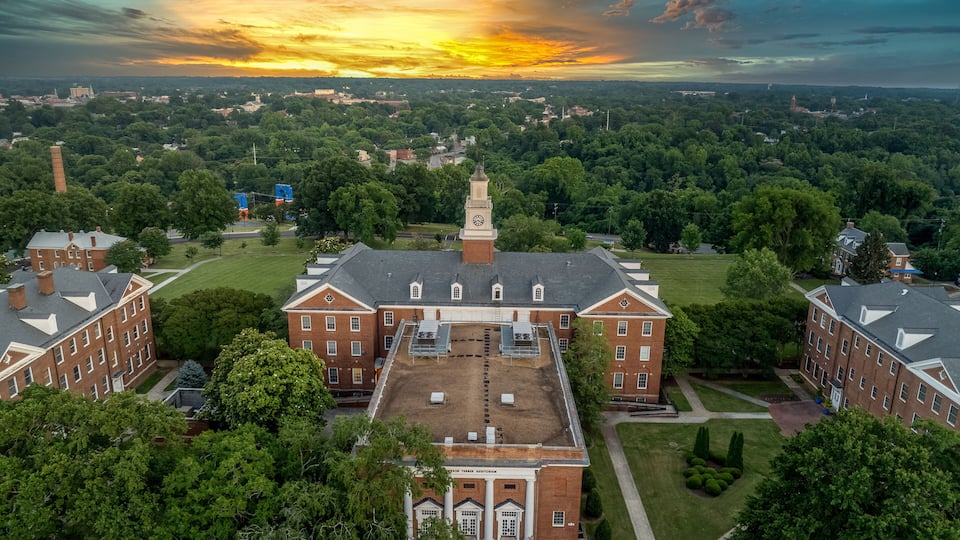 A large brick building with a clock tower, surrounded by green trees and a sunset sky.