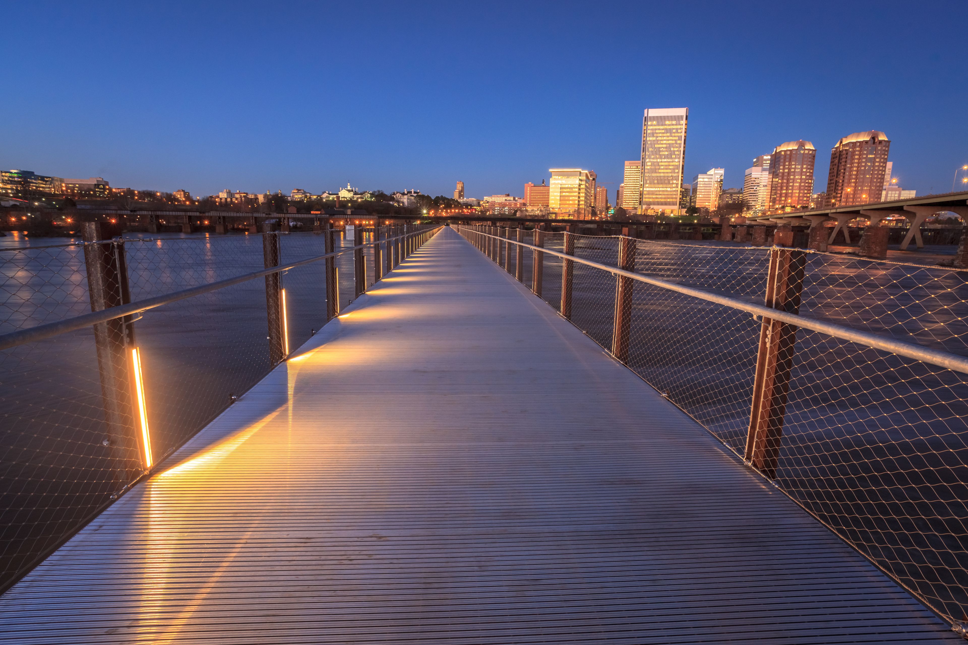 A waterfront promenade with a metal railing, reflecting lights from the city skyline in the background.