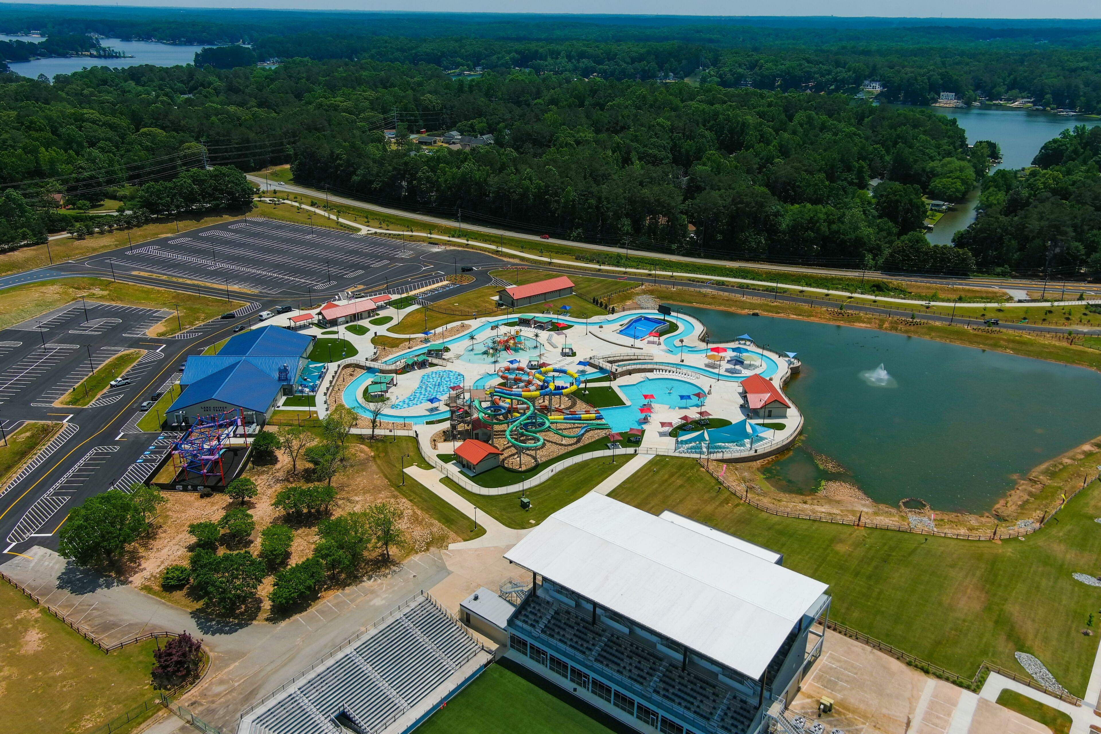 a stunning aerial shot of long winding pools with blue water and colorful waterslides with a lake with a water fountain surrounded by lush green trees and grass at Lake Spivey Recreation Center