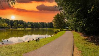 A serene park with a pond, a walking path, and a sunset in the background.