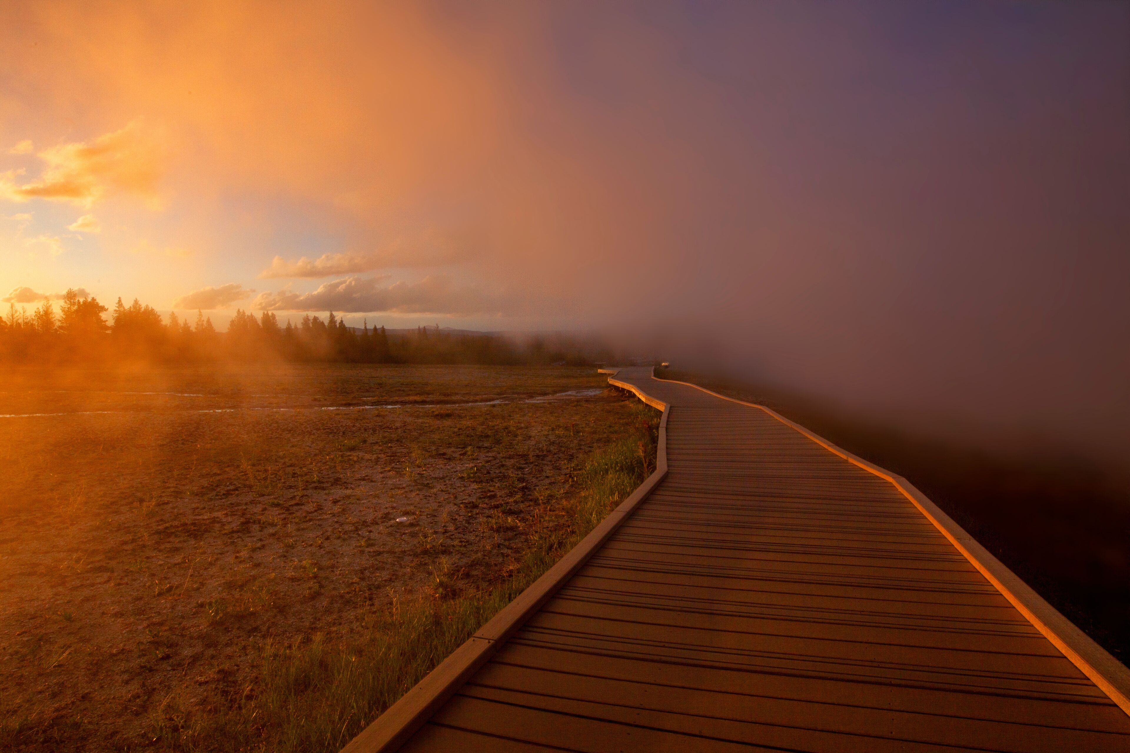 Un paseo marítimo de madera que se adentra en un paisaje brumoso, con una puesta de sol al fondo.
