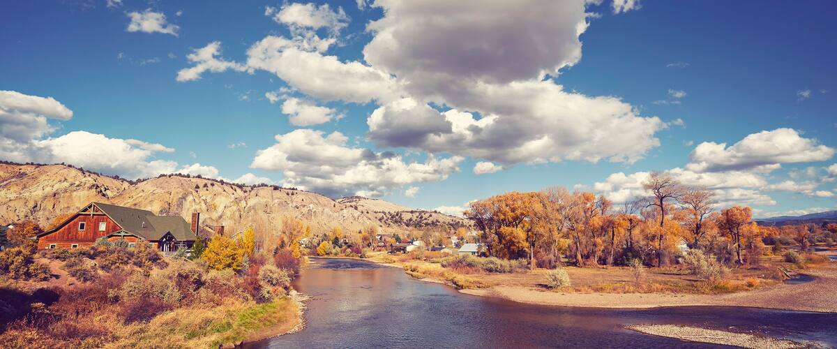 A river flowing through a valley with a red barn and houses, surrounded by autumn-colored trees and a mountainous backdrop.