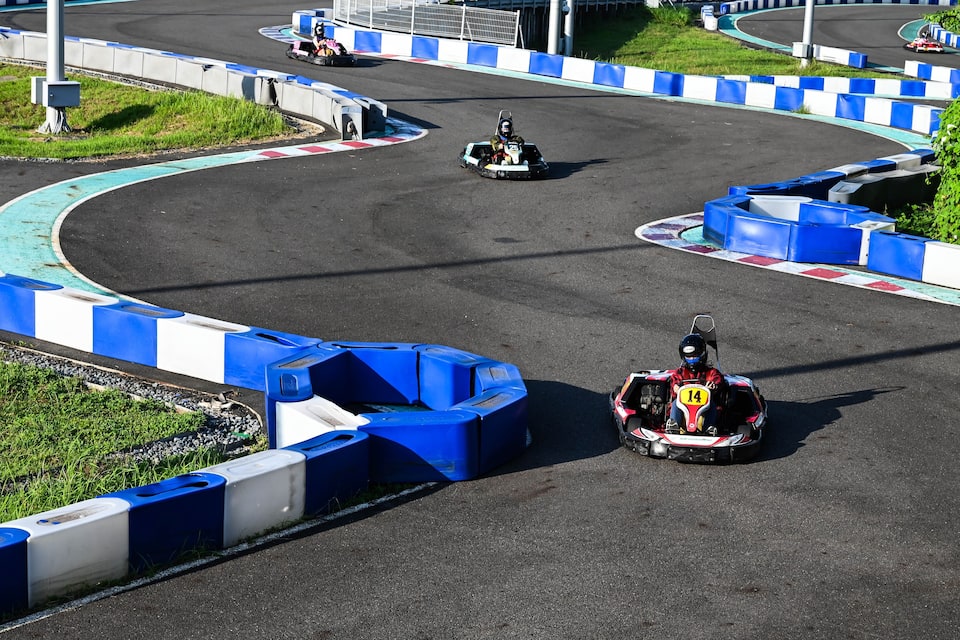 Pista de corrida de kart com barreiras azuis e brancas, um piloto com macacão vermelho e o número 14, e outro kart ao fundo.