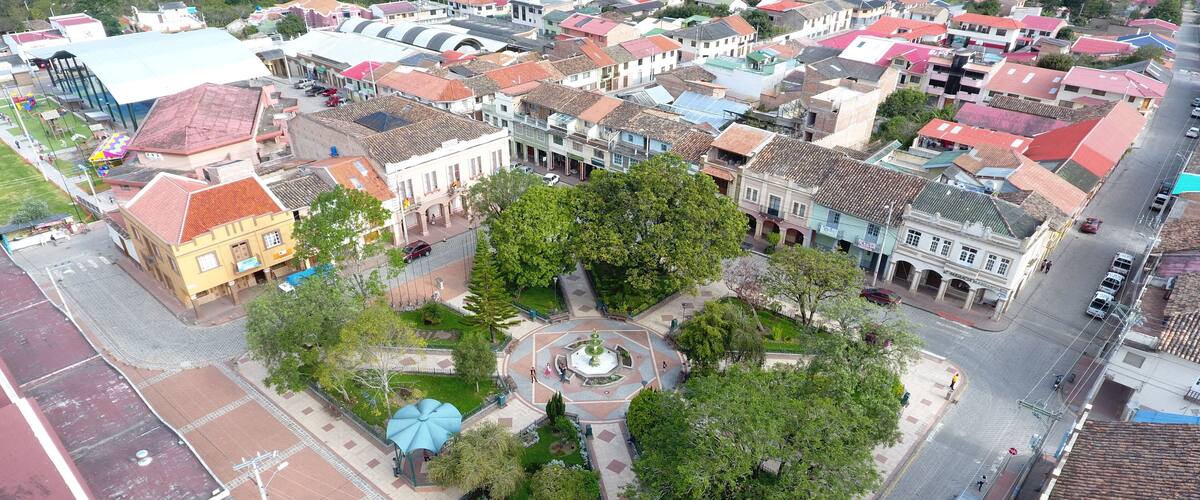 An aerial view of a town square with a central fountain, surrounded by trees and buildings with red and white roofs.