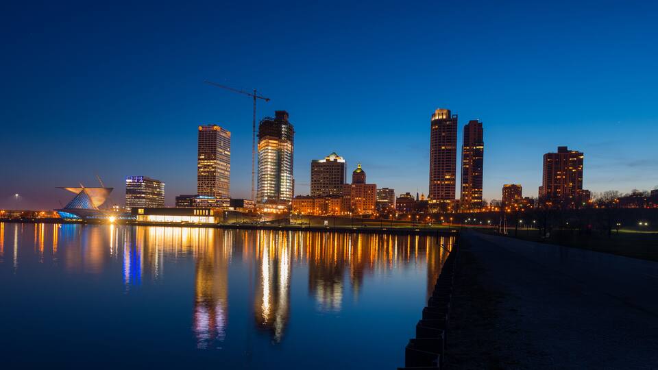 A city skyline at dusk with illuminated buildings reflecting on the water.