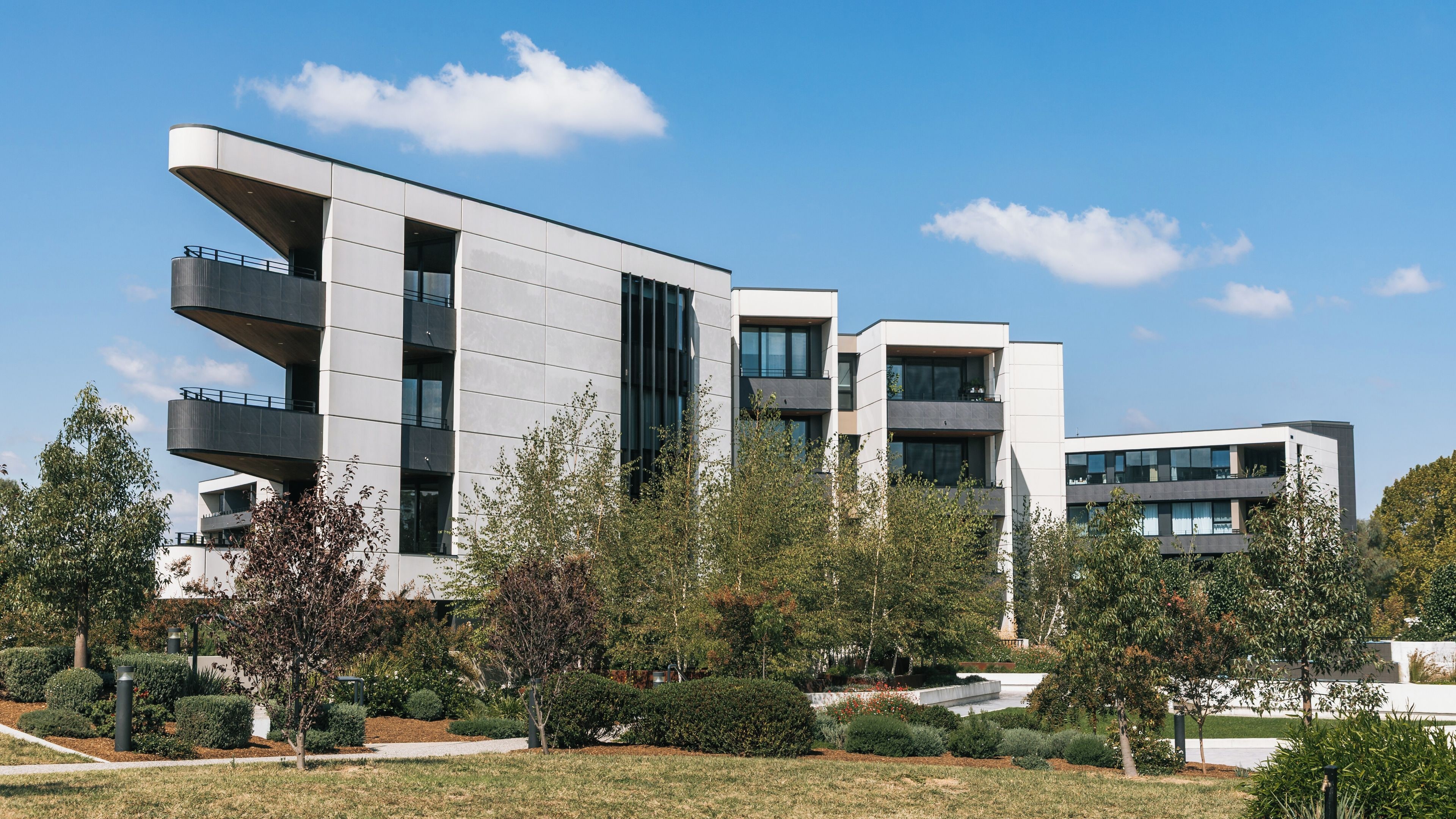 A modern multi-story building with balconies, surrounded by trees and a clear blue sky.