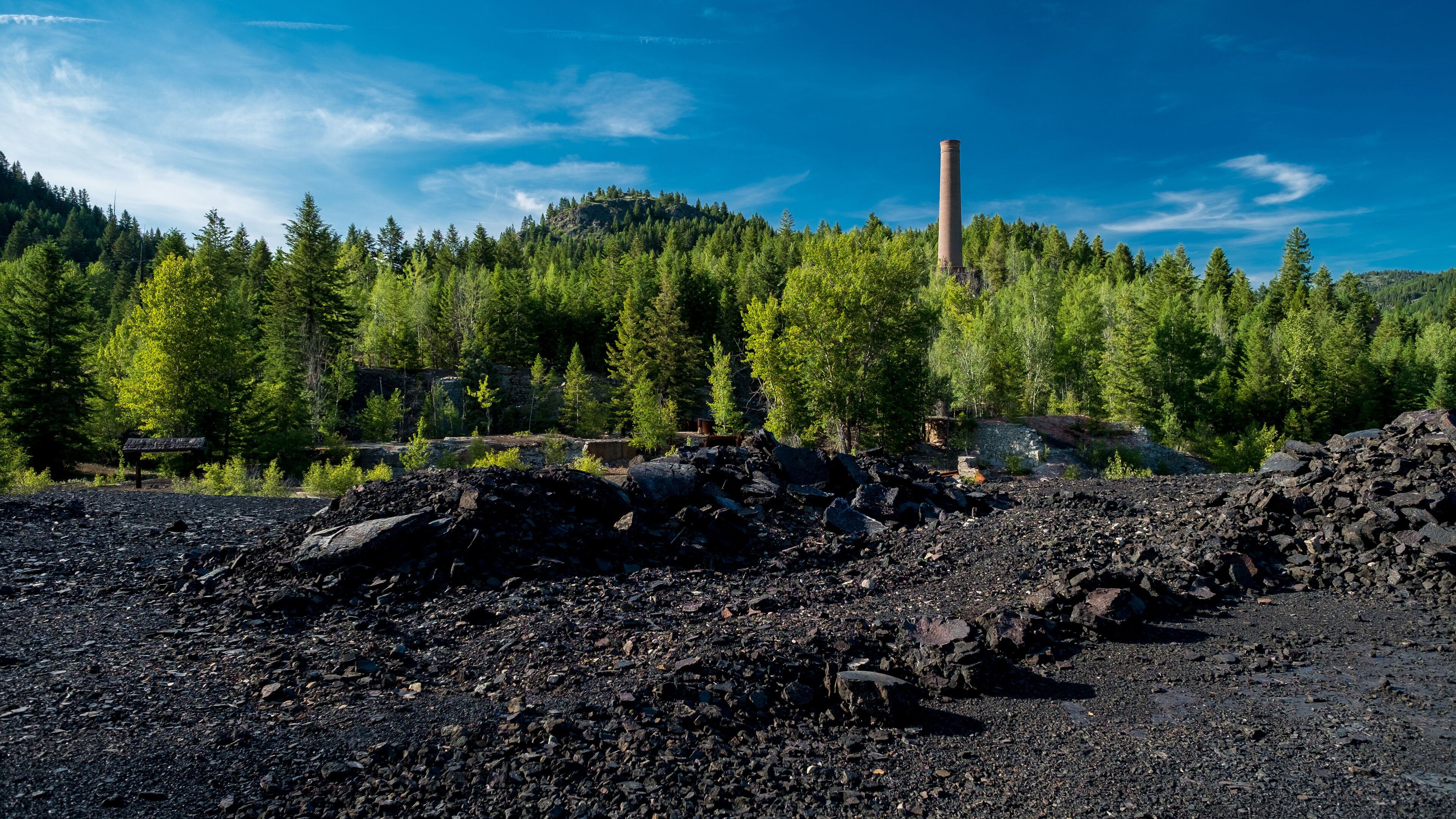 A rocky terrain with a forested hill and a tall chimney in the background.