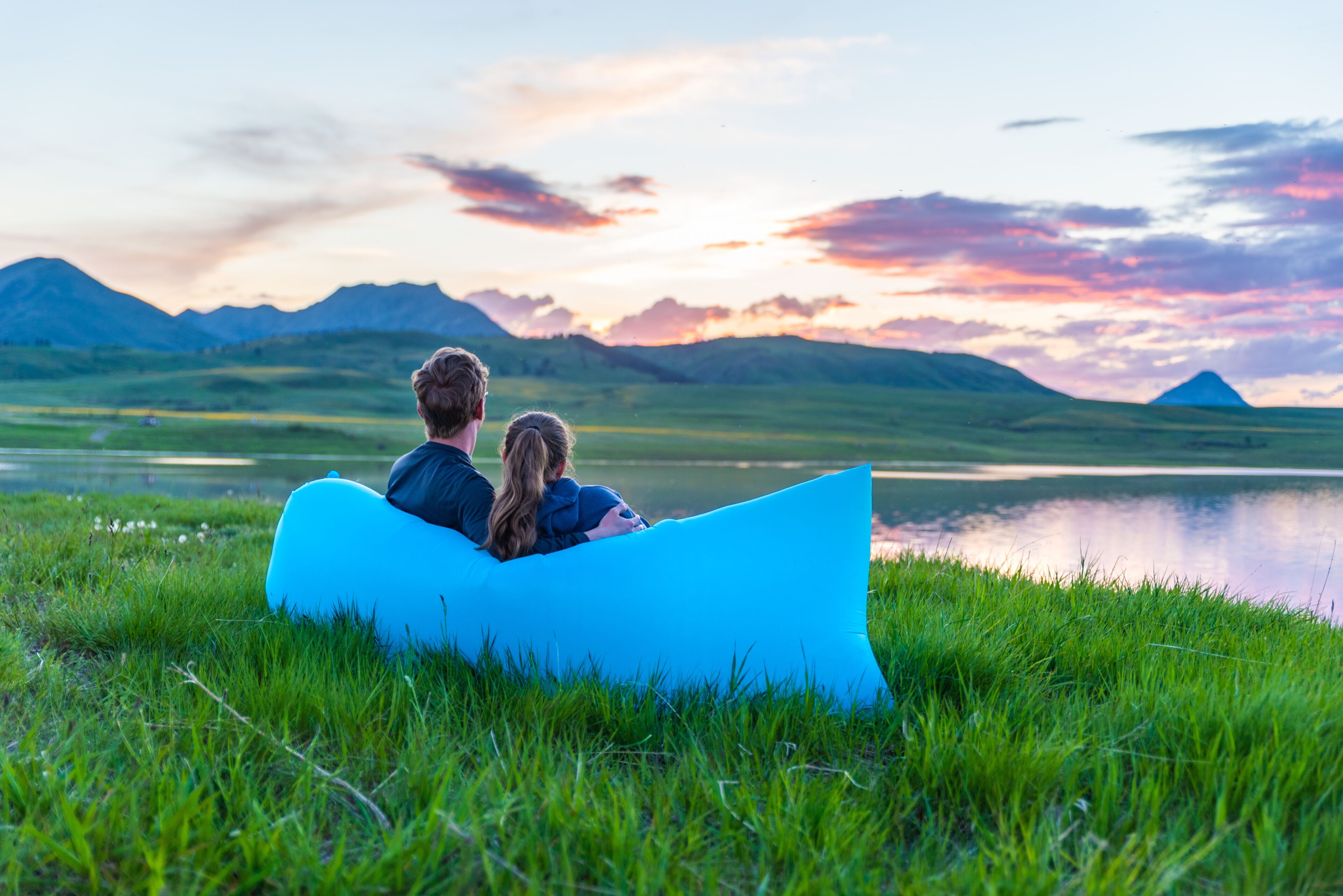 A couple sitting on a blue inflatable raft by a lake with mountains in the background.