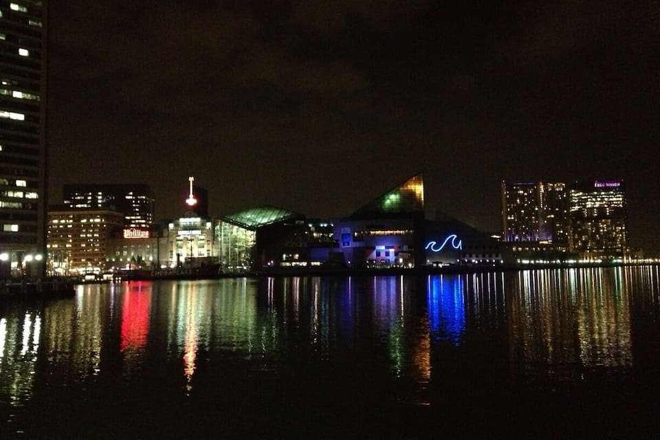 Une vue nocturne d’une ville, avec des bâtiments illuminés dont les reflets se mirent dans l’eau.