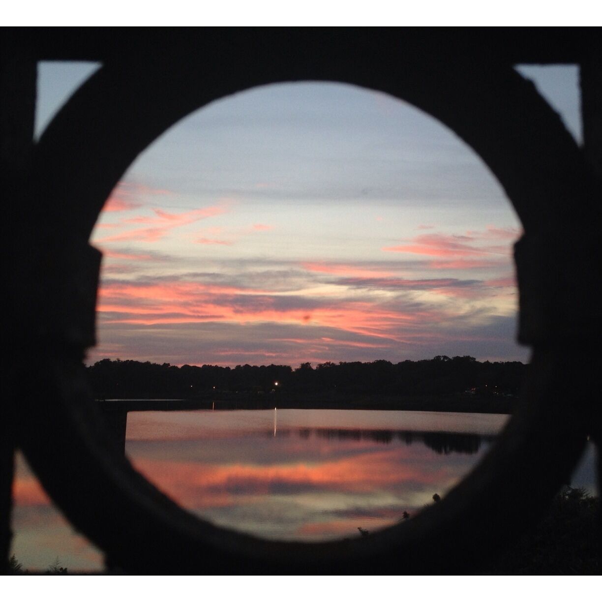 This beautiful Baltimore  sunset is peeking out through a gap in the fence at Druid Hill Park. The reflection on the lake is  mesmerizing--but don't stay too long past dusk, because it's not the safest area. NOTE: this is a completely unaltered iPhone photo