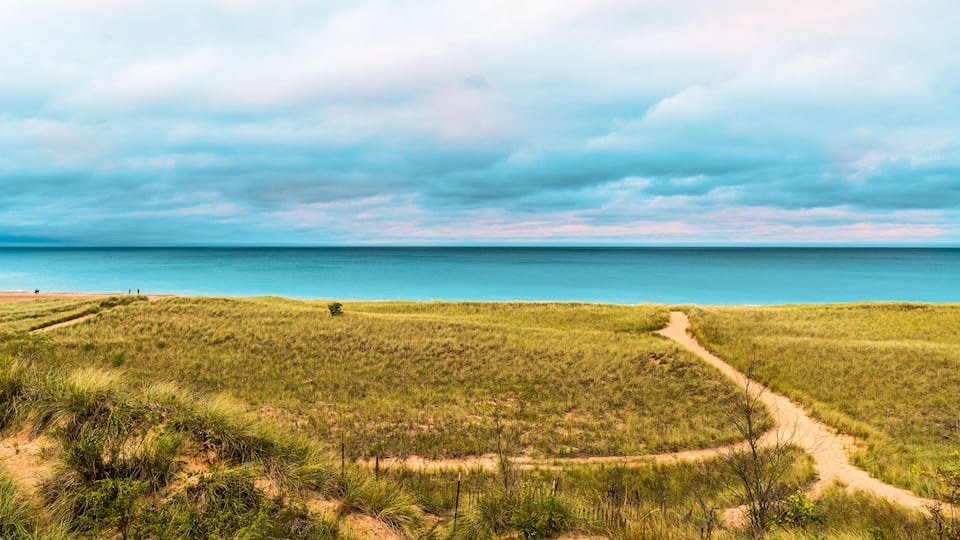 A coastal landscape with a sandy beach, grassy dunes, and a clear view of the ocean under a cloudy sky.