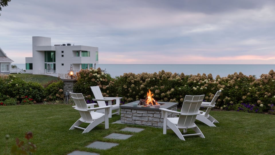 A modern house with a stone fire pit and white Adirondack chairs in a garden with a view of the ocean.
