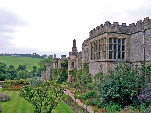 A historic stone building with large windows and a turret, surrounded by lush gardens and green fields.