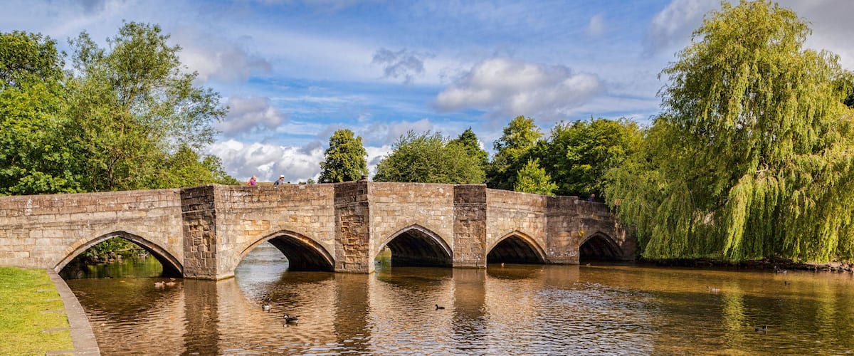 A stone bridge with multiple arches over a calm river, surrounded by lush greenery and trees.