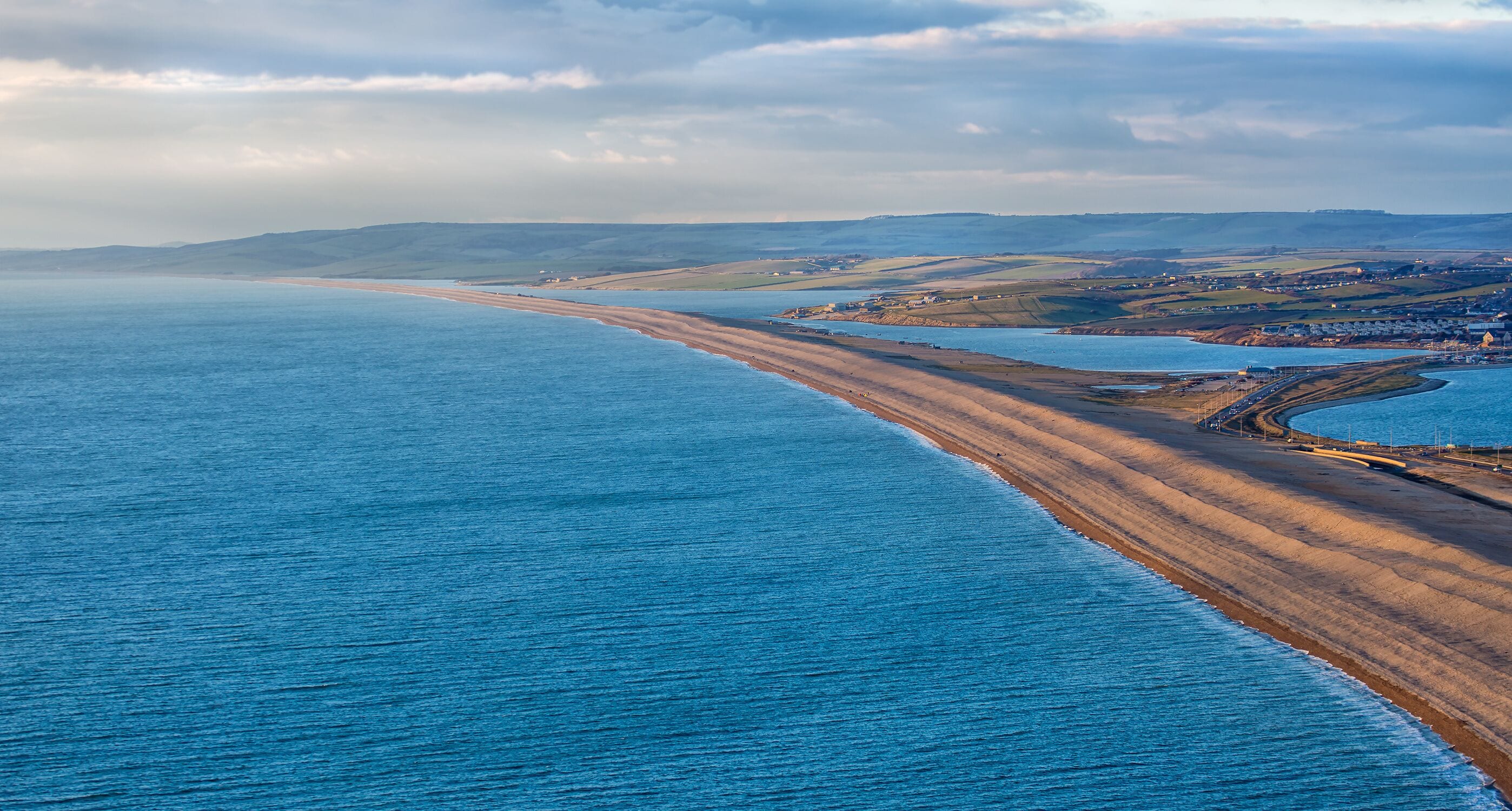 Un paisaje costero con una playa extensa, un mar tranquilo y colinas onduladas al fondo.