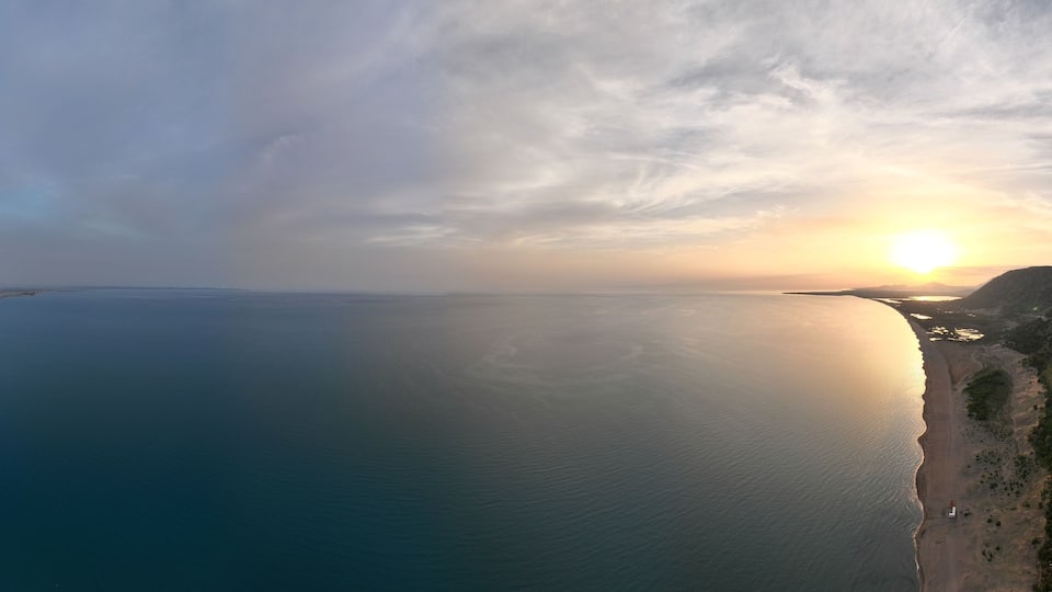 A coastal landscape with a calm sea, a clear sky, and a setting sun.