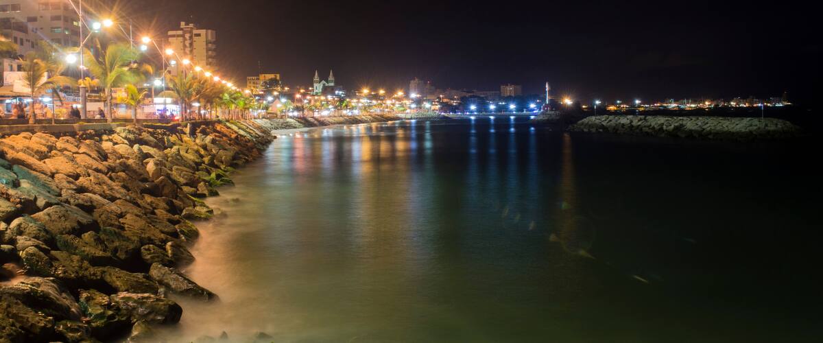 A nighttime coastal cityscape with a rocky shoreline, illuminated streets, and distant buildings.