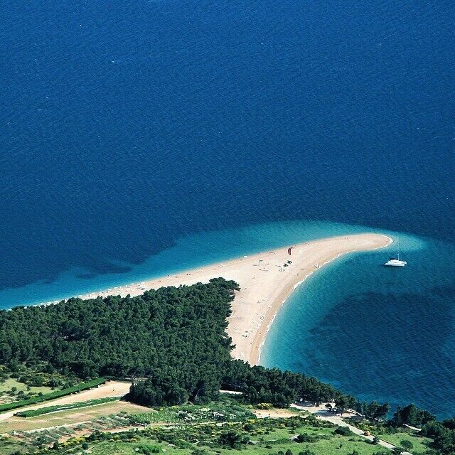 An aerial view of a coastal area with a curved beach, dense forest, and a boat in the water.