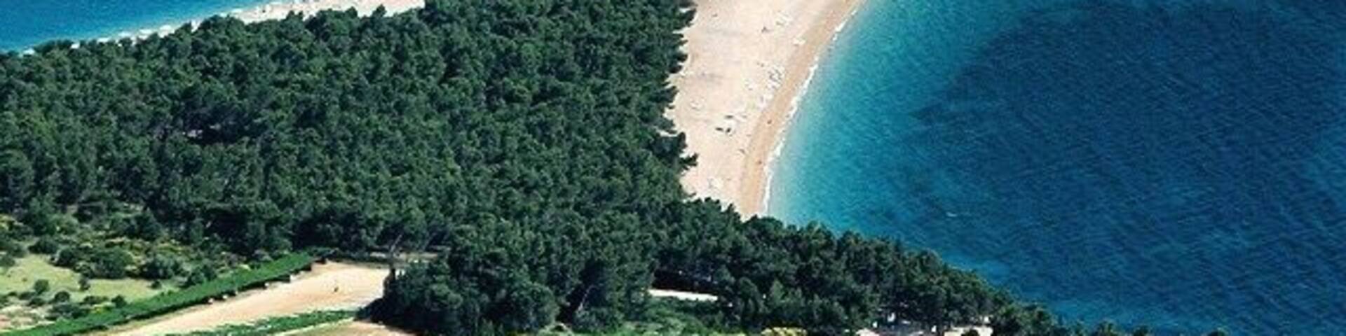 An aerial view of a coastal area with a curved beach, dense forest, and a boat in the water.