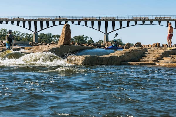 A river with a bridge, kayakers, and people standing on rocks.
