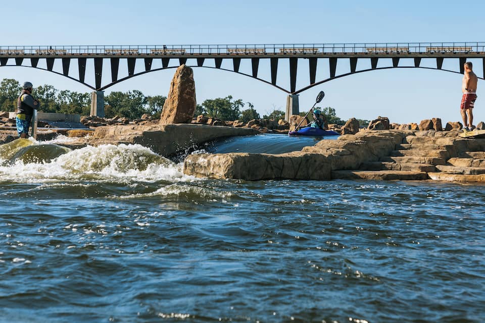 A river with a bridge, kayakers, and people standing on rocks.