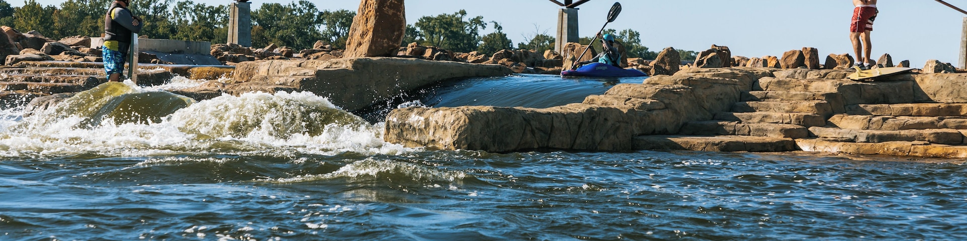 A river with a bridge, kayakers, and people standing on rocks.