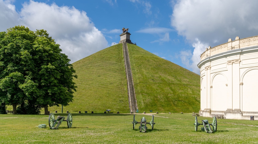 A grassy hill with a statue on top, a large tree, and a white building with columns.