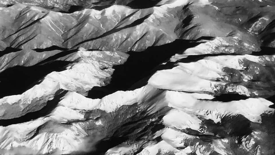 A black and white aerial view of rugged mountain peaks with snow-covered slopes and deep valleys.