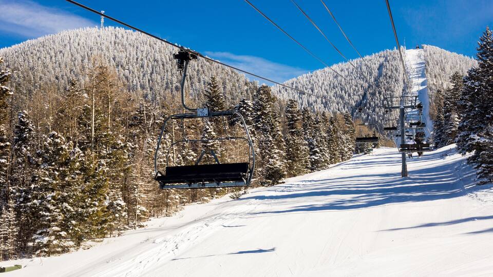 A ski lift on a snowy mountain with trees and a clear blue sky.