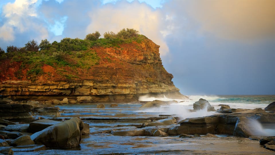 A coastal landscape with a rocky cliff, waves crashing against the shore, and a cloudy sky.