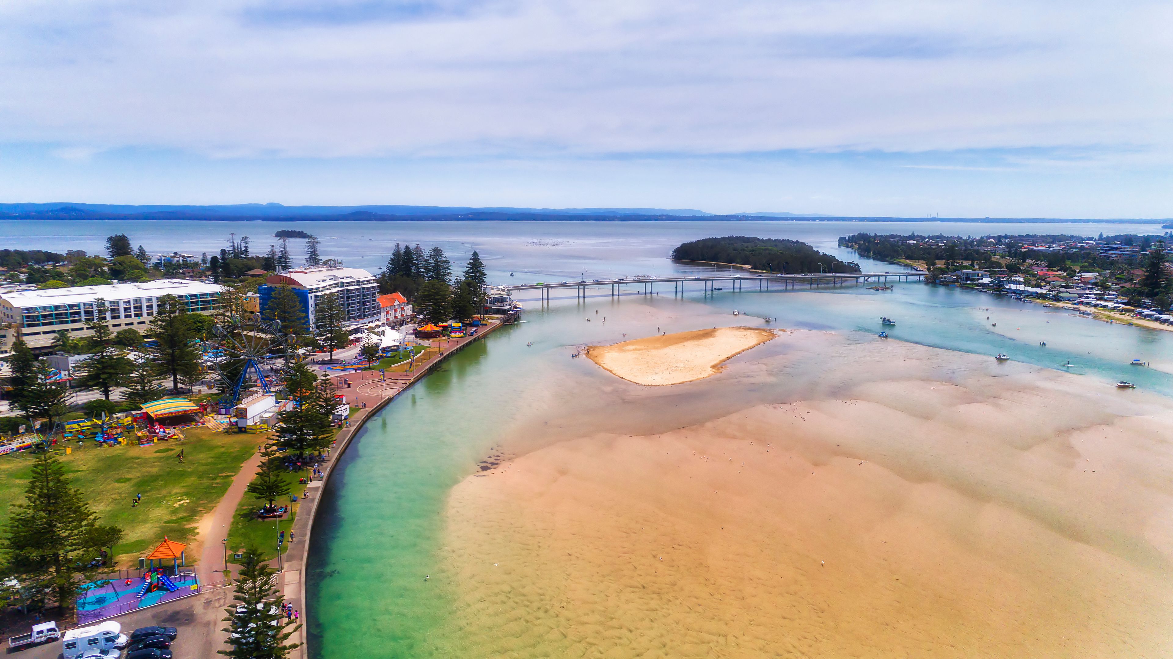 A coastal area with a sandy beach, a river, a bridge, and a park with playgrounds.