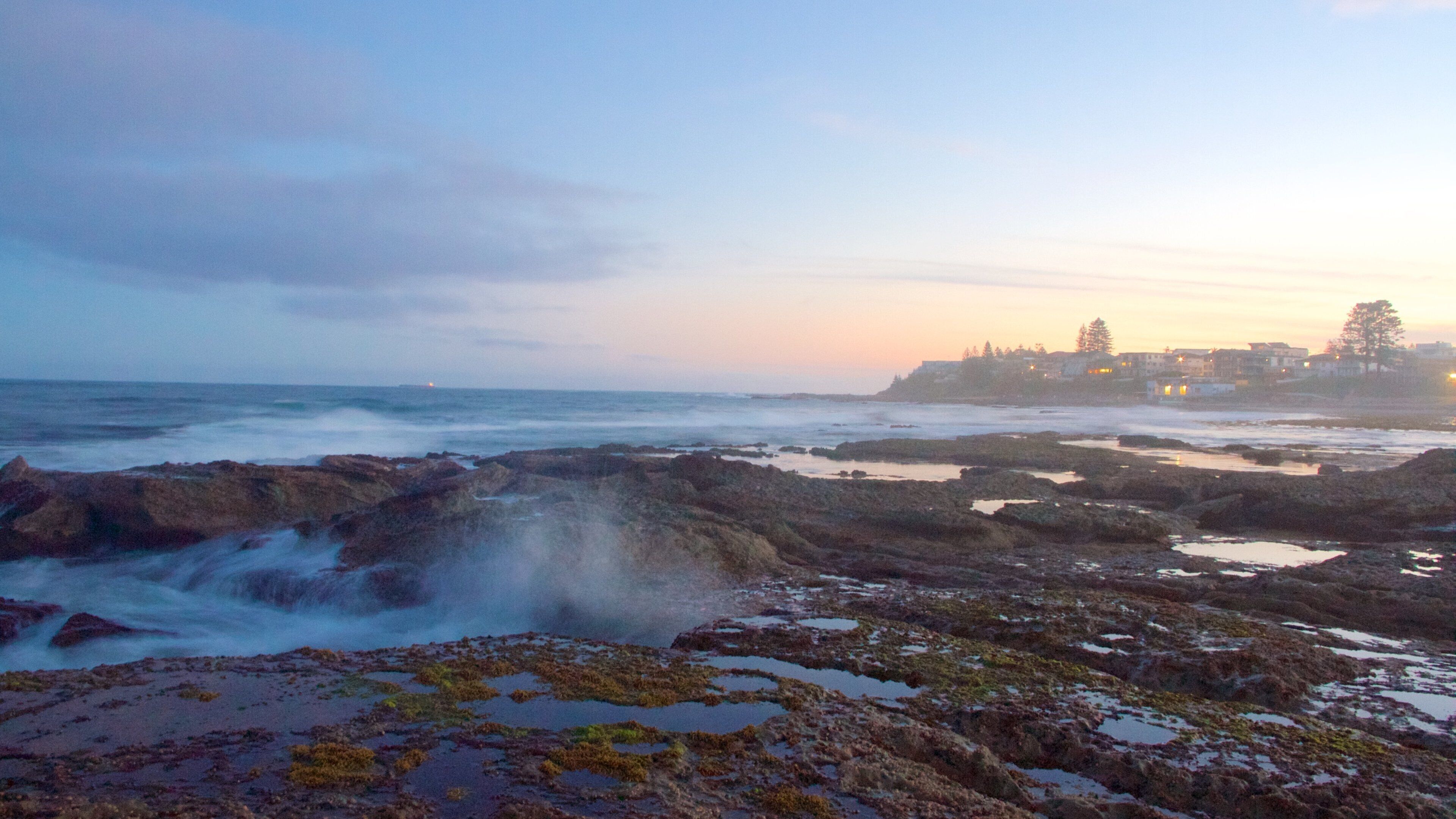 A rocky shoreline with tide pools and a distant cityscape at sunset.