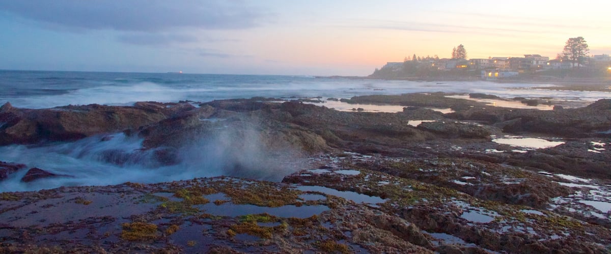 A rocky shoreline with tide pools and a distant cityscape at sunset.