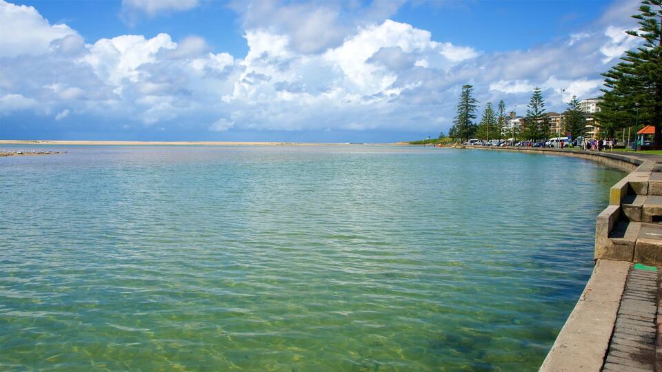 A clear waterfront with a sandy beach, a concrete walkway, and buildings in the background.
