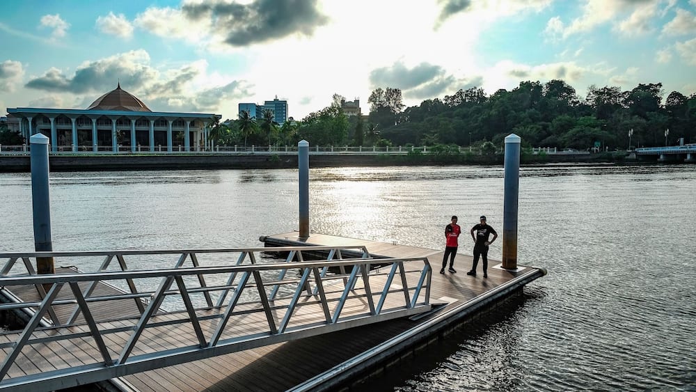 Eine Holzstege streckt sich in einen ruhigen Fluss, am Ende stehen zwei Personen. Im Hintergrund ist ein großes Gebäude mit einer Kuppel und einer Brücke über dem Fluss zu sehen.