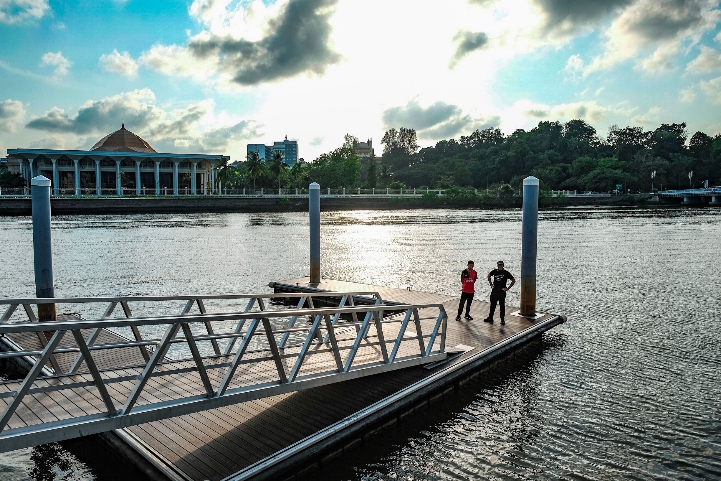 A wooden dock extending into a calm river, with two people standing at the end. In the background, there is a large building with a dome and a bridge over the river.