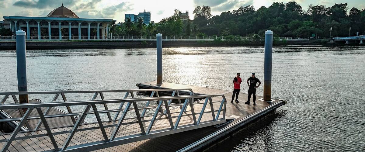 A wooden dock extending into a calm river, with two people standing at the end. In the background, there is a large building with a dome and a bridge over the river.