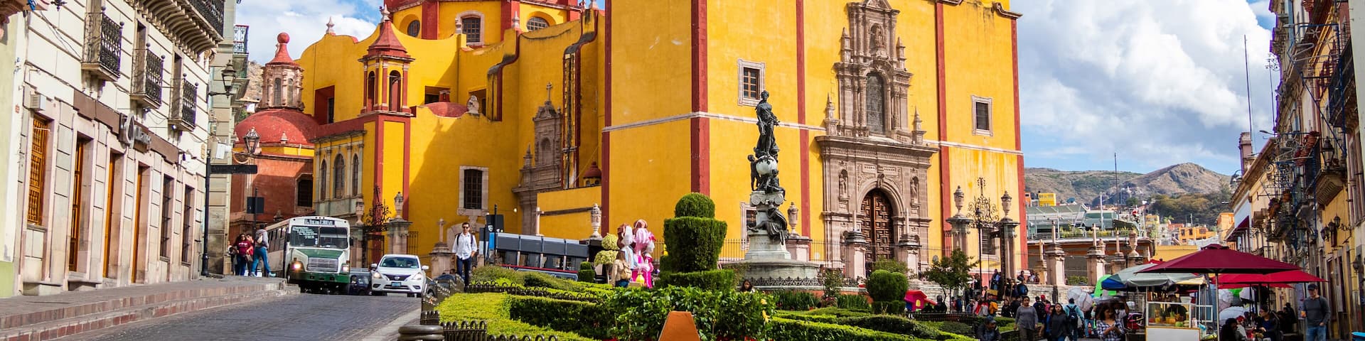 A historic yellow church with a red dome and bell towers, surrounded by a cobblestone plaza with a fountain and people.