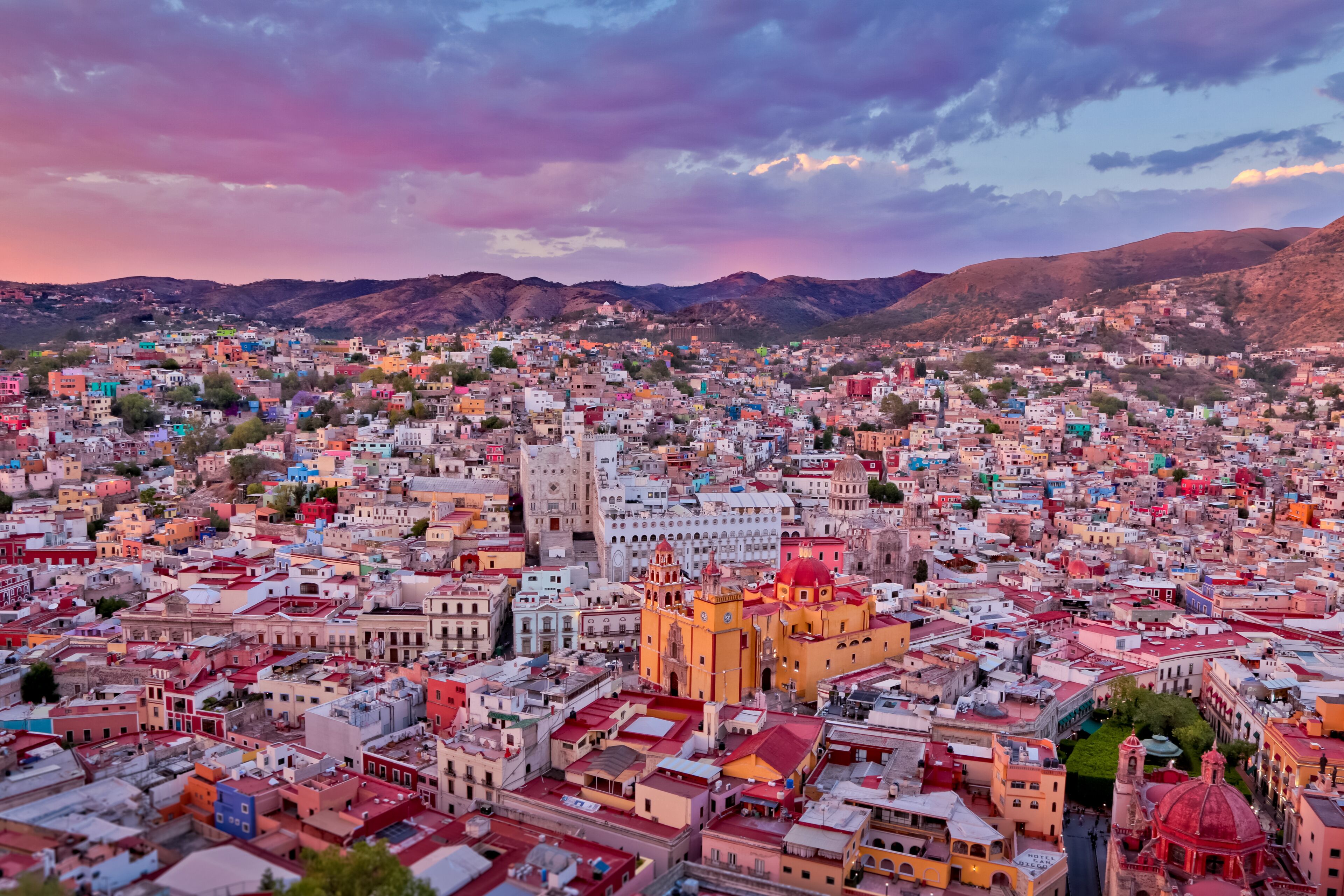 A densely populated cityscape with colorful buildings, a prominent church with a red dome, and mountains in the background.