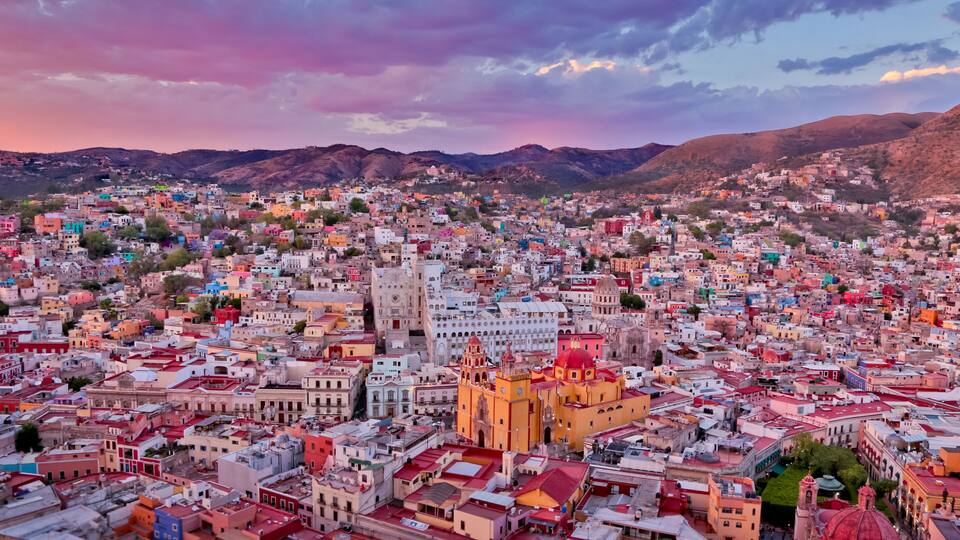 A densely populated cityscape with colorful buildings, a prominent church with a red dome, and mountains in the background.