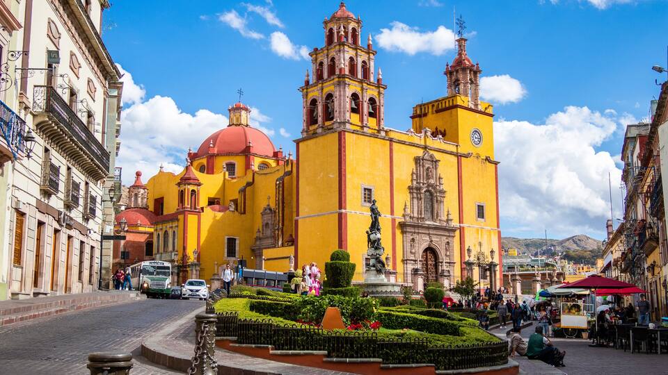 A historic yellow church with a red dome and bell towers, surrounded by a cobblestone plaza with a fountain and people.