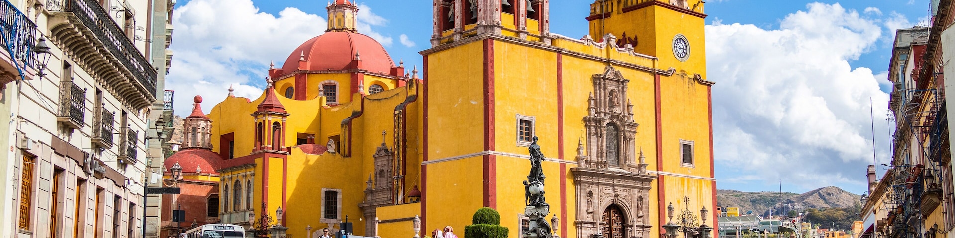 A historic yellow church with a red dome and bell towers, surrounded by a cobblestone plaza with a fountain and people.
