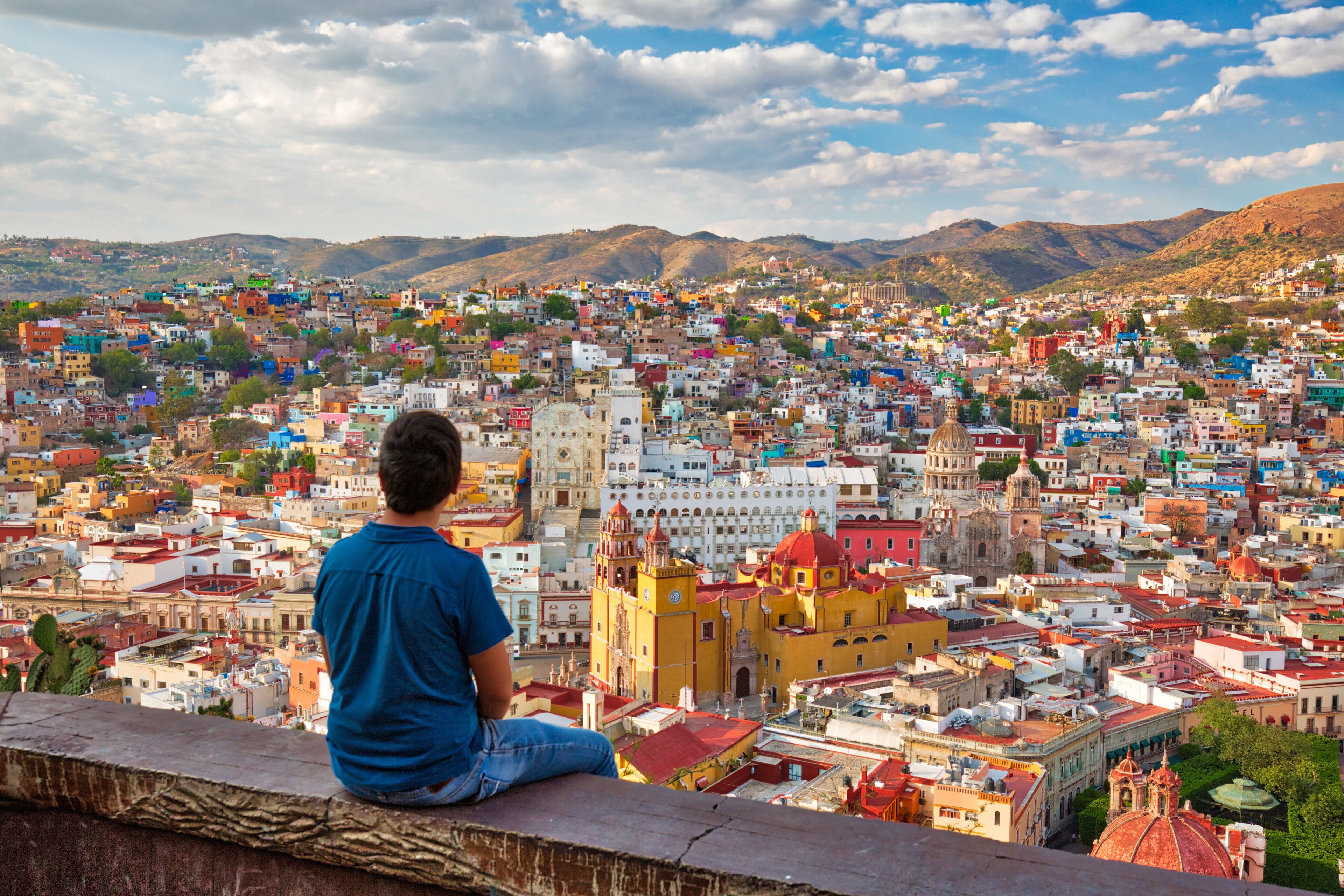 A person sitting on a ledge overlooking a densely packed cityscape with colorful buildings and a prominent church with a red dome.