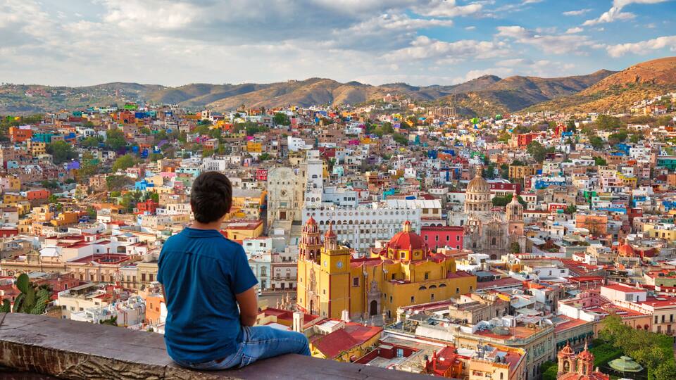 A person sitting on a ledge overlooking a densely packed cityscape with colorful buildings and a prominent church with a red dome.