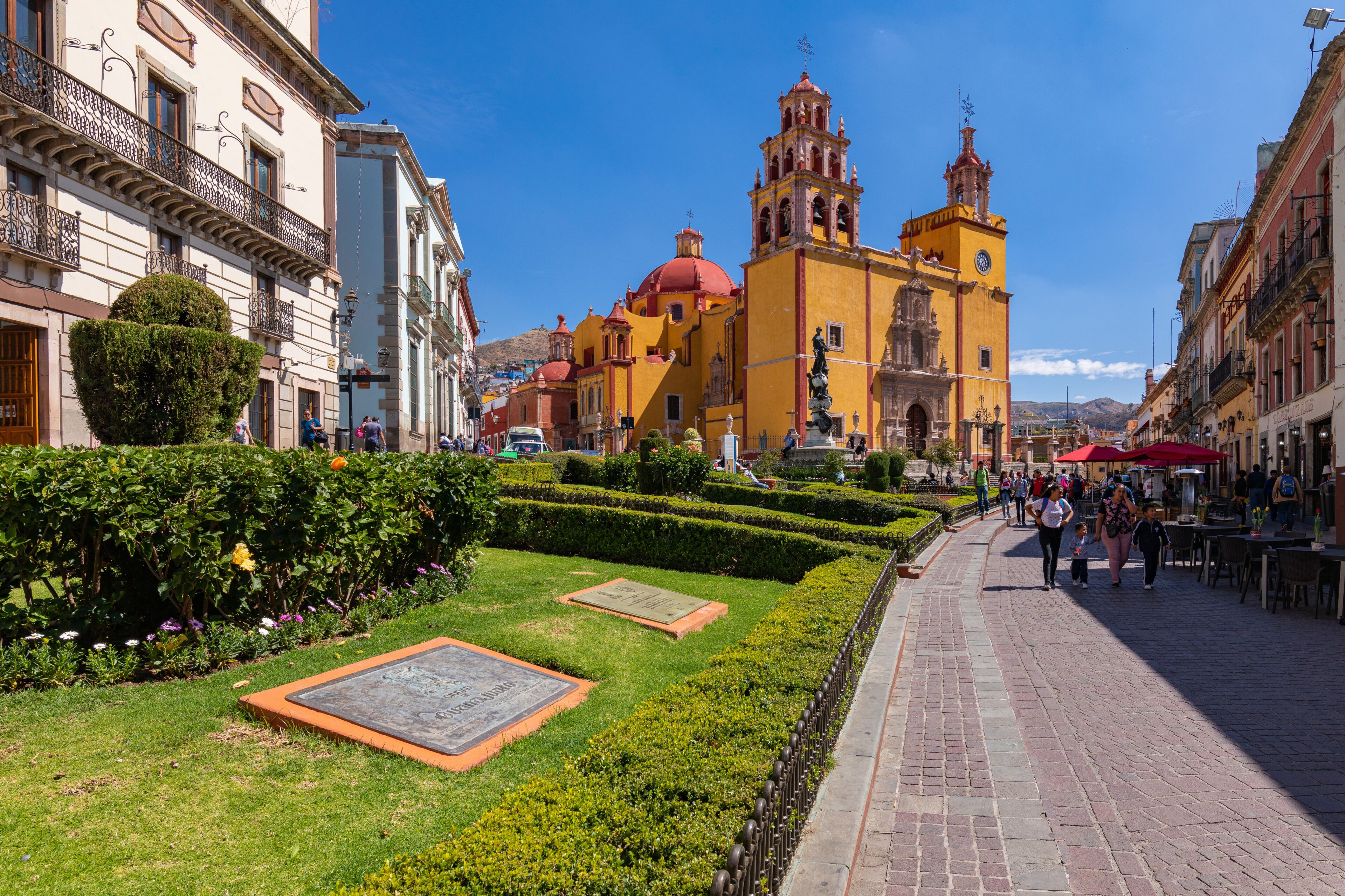 Una plaza histórica con una iglesia amarilla destacada, jardines cuidados y calles adoquinadas.