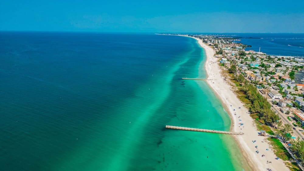 Eine Küstenstadt mit einer langen Pier, die sich in das türkisfarbene Meer erstreckt, einem Sandstrand und Wohngebäuden entlang der Uferlinie.